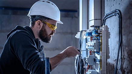 Mężczyzna (elektryk) w białym kasku, żółtych okularach i czarnej bluzie ze śrubokrętem w ręce sprawdza instalację elektryczną w skrzynce z bezpiecznikami.